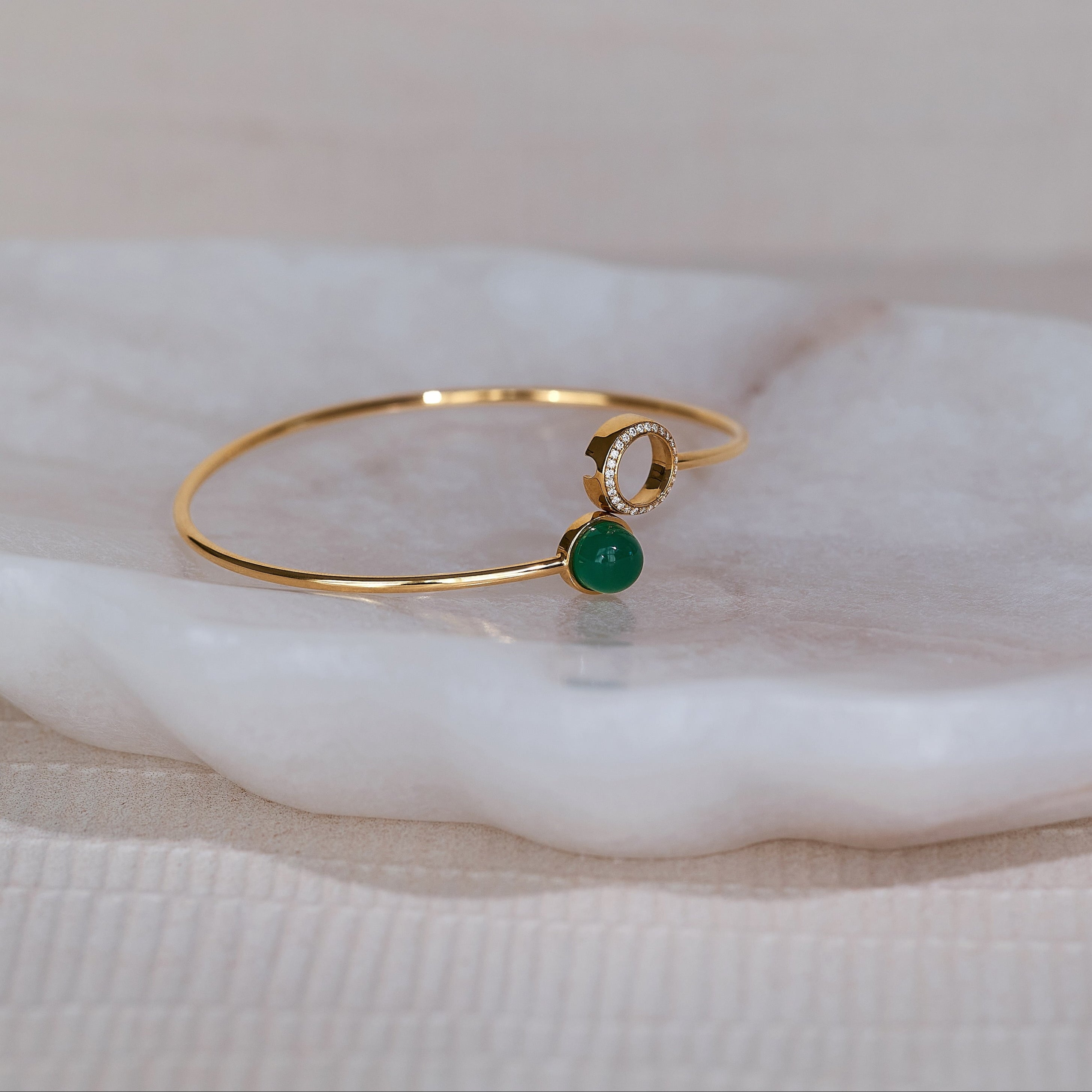 A yellow gold bracelet with a bezel-set green onyx center stone and a diamond halo, displayed on a white marble surface.