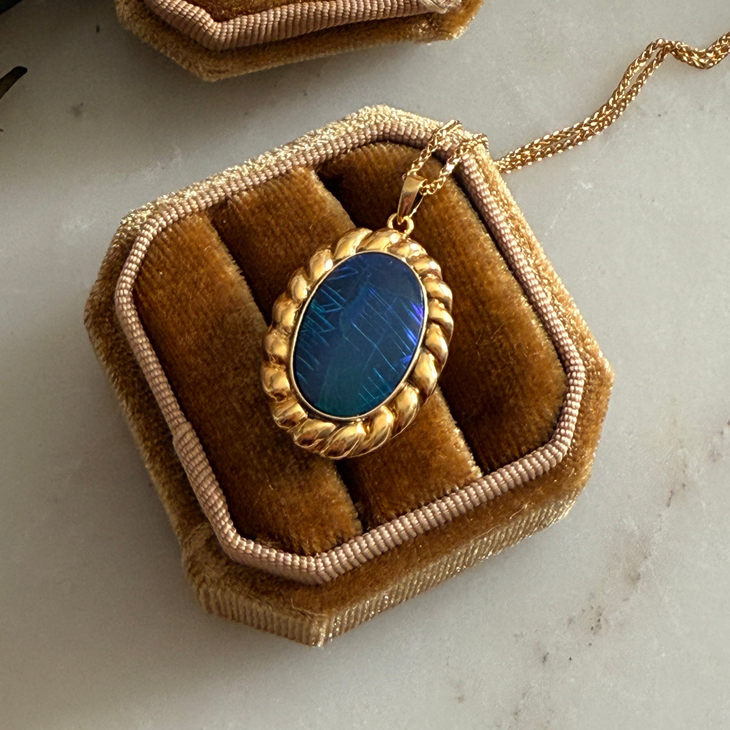 Gold necklace with indigo opal stone pendant on a brown velvet stand, surrounded by flowers.
