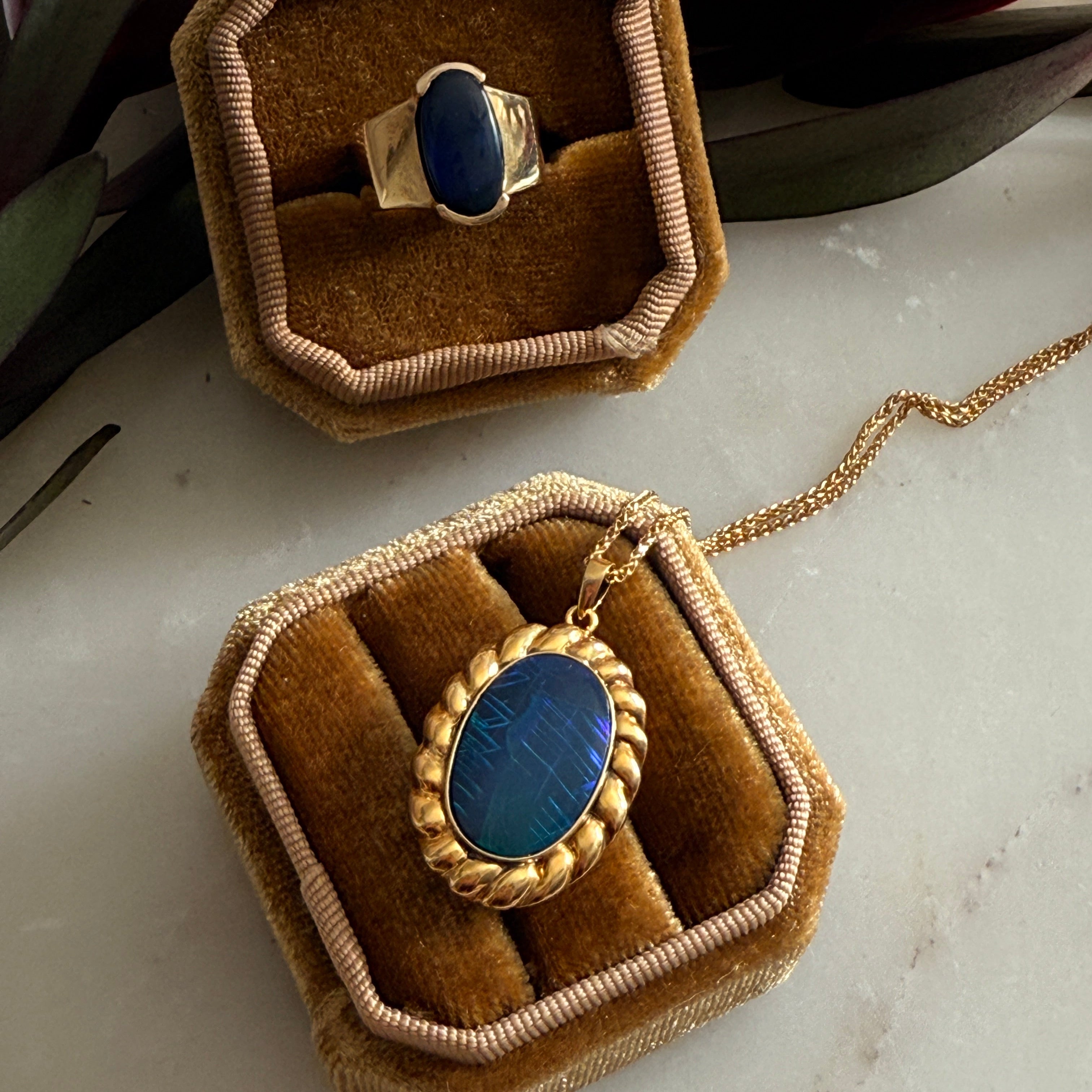 Gold necklace with indigo stone pendant on a brown velvet stand, surrounded by flowers.