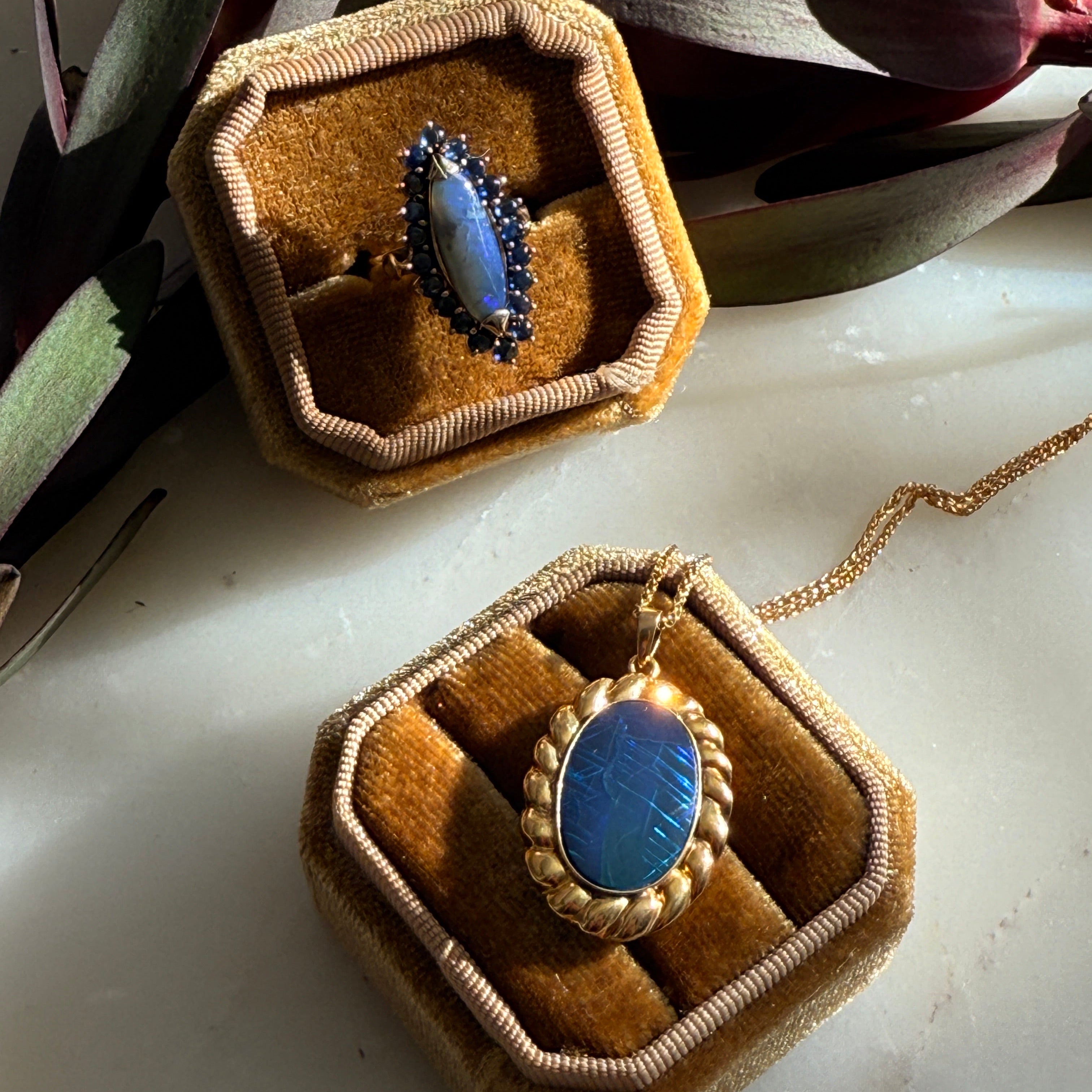 Two gold rings with blue fire opal  stones in hexagonal brown boxes on a light surface.